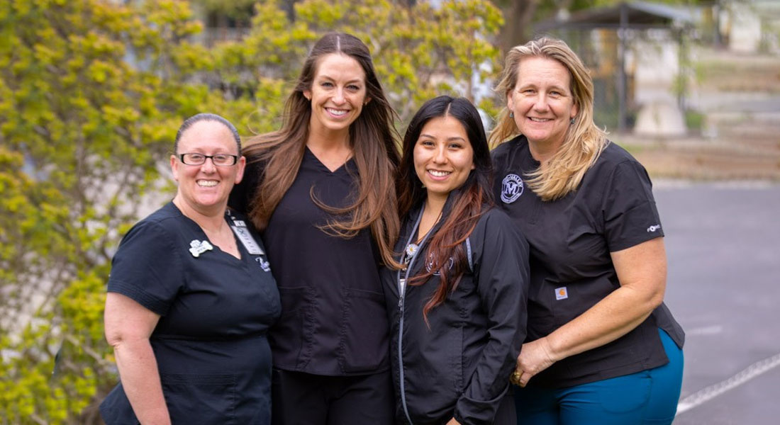 Four women wearing black scrubs, possibly veterinarians, standing closely together, smiling at the camera in an outdoor setting with greenery and a building in the background.