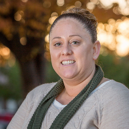 A woman with light skin and light brown hair pulled back in a bun is smiling at the camera. She is wearing a beige sweater and a green knitted scarf, looking every bit the dedicated veterinarian she is. The background shows blurred trees and a warm, golden sunset.