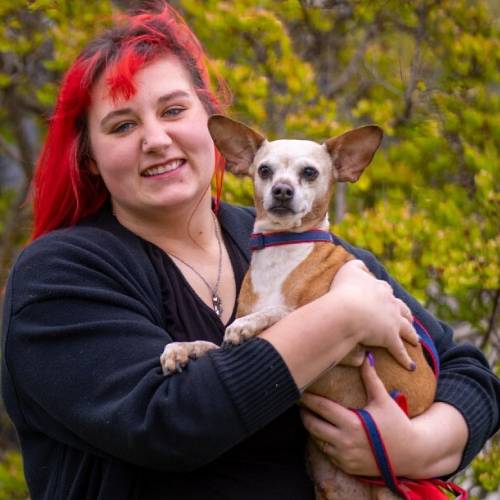 A person with vibrant red hair smiles while holding a small, tan and white dog with large ears. They are outdoors with green foliage in the background, appearing content and happy. The scene evokes the warmth of a veterinarian bonding with their cherished companion.