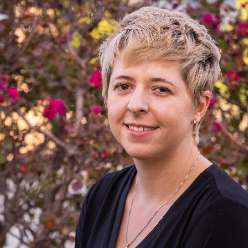 A person with short blond hair, who is a veterinarian, smiles while standing outdoors in front of a bush with pink flowers. They are wearing a black top and a silver necklace. The background includes blurred leaves and blossoms.