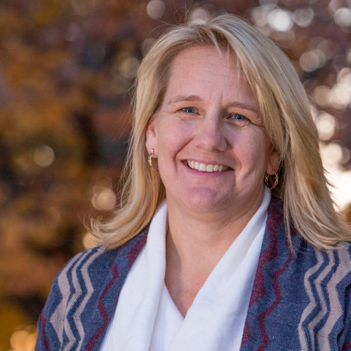 A blonde woman, who happens to be a veterinarian, smiles at the camera. She is wearing a patterned jacket and a white scarf. The background is blurred with autumnal colored leaves.