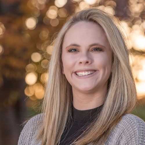A blonde woman with straight hair smiles widely at the camera. She is wearing a dark top with a light-colored cardigan. The background is blurred with warm tones and soft bokeh effects, suggesting a sunset or early evening setting. The cheerful vet seems to exude warmth and compassion.