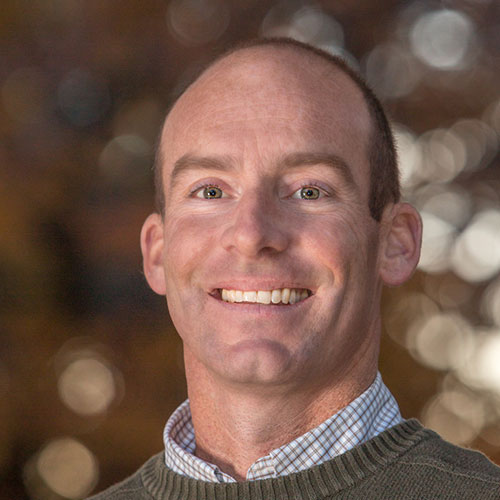 A smiling man with a shaved head and green sweater over a collared shirt stands against a blurred background with warm, bokeh lighting. The experienced veterinarian radiates warmth, capturing the essence of compassionate care.