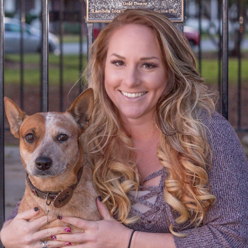 A woman with long, wavy blonde hair, perhaps a veterinarian, smiles at the camera while holding a brown and white dog. They are outside in front of a black metal fence, with a green park visible in the background. She is wearing a purple sweater and has manicured nails.