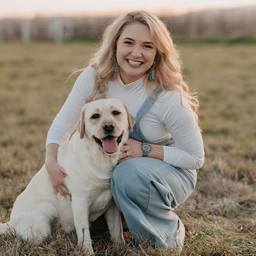 A woman with long blonde hair, wearing a white top and light blue overalls, kneels in a grassy field while smiling and hugging a happy yellow Labrador Retriever.