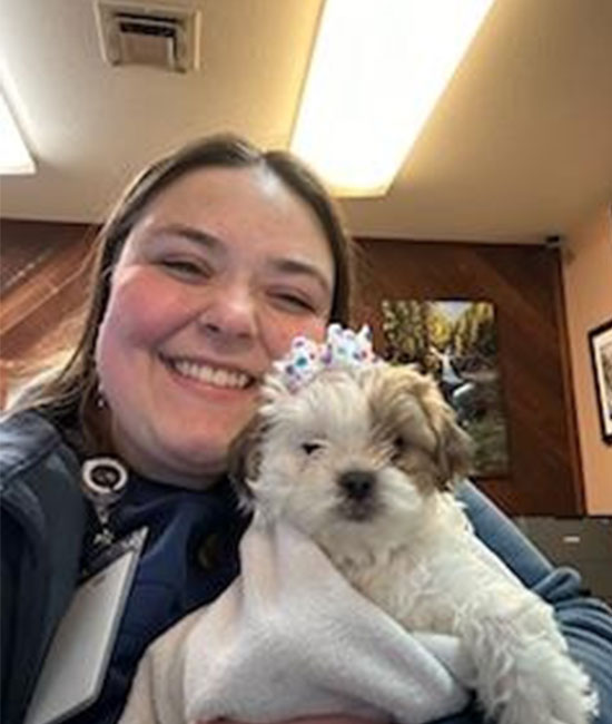 A woman smiling and holding a small, fluffy puppy wrapped in a white blanket. The puppy is wearing a cute headband with colorful bows. They appear to be indoors, possibly at the vet or a clinic, with fluorescent lights overhead and pictures on the walls in the background.