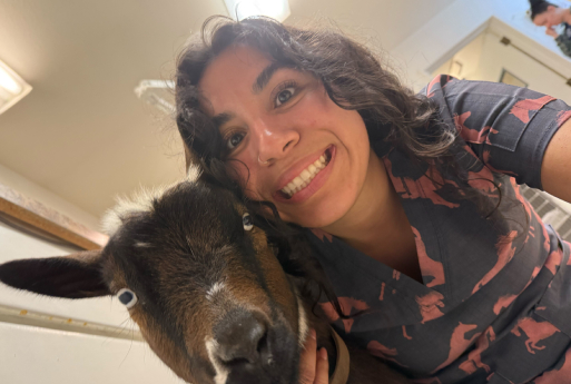 A smiling woman with curly hair takes a close-up selfie indoors with a brown and white goat, both looking at the camera.
